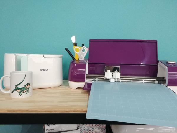 A Cricut Mugpress and a Cricut on a table surrounded by craft supplies.