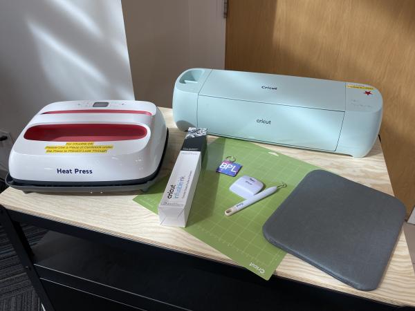 A Cricut Heat Press and a Cricut on a table surrounded by craft supplies.