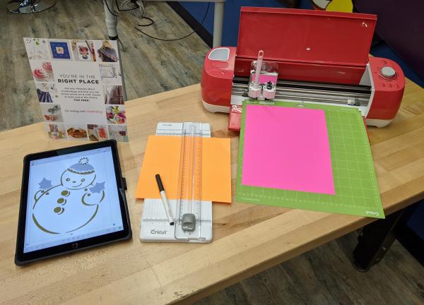 A table holding a tablet with a picture of a snowman on the screen. Next to the tablet is craft paper bring fed into a Cricut.